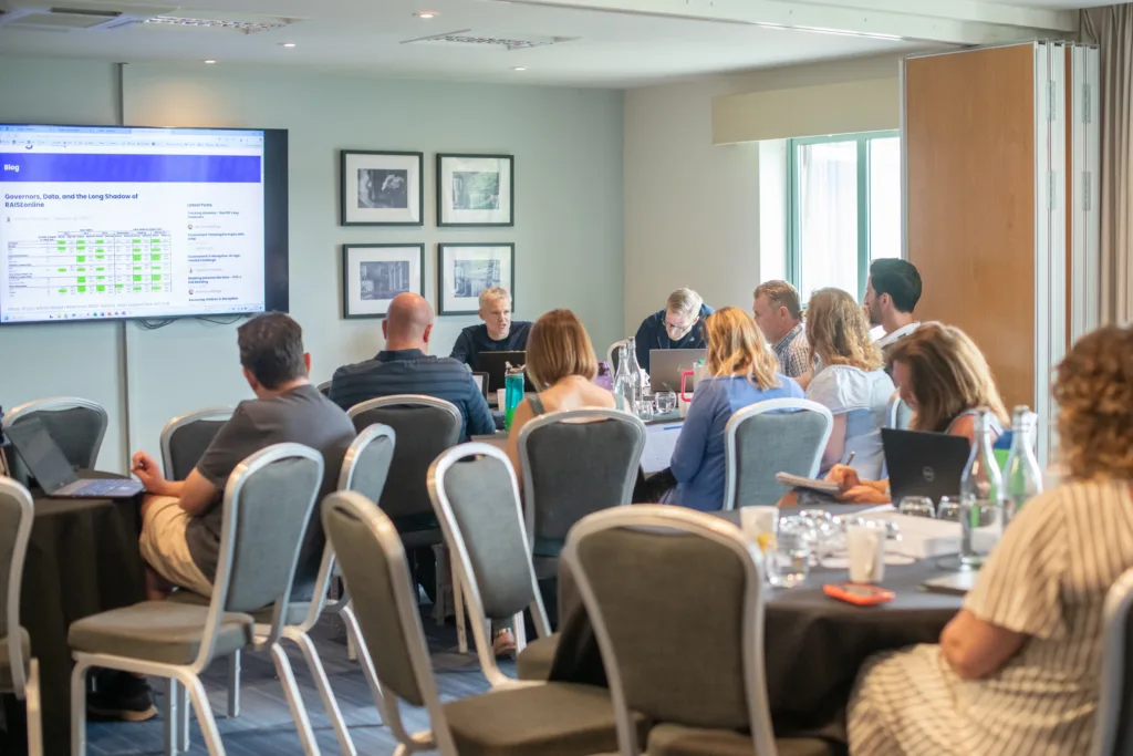 A conference room showing a Databusters session in progress. Guests are sitting around tables listening to James Pembroke speak and present on a projector board. 